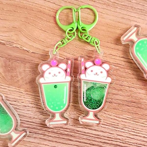 May include: Clear acrylic keychains featuring a cartoon bear in a green drink with a pink straw and cherry. The keychains have a green glitter filling and are attached to a green clasp. The background is a light brown wooden surface.