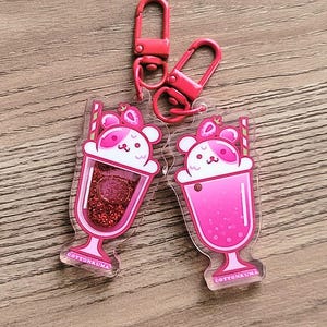 May include: Two acrylic keychains featuring a pink milkshake design with a bear face, strawberries, and a straw. The keychains have red lobster clasp attachments. The text "COTTONKUMA" is at the bottom of each keychain.