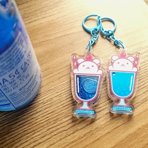 May include: Two acrylic keychains featuring a cartoon bear in a milkshake glass design. The keychains have blue accents, pink details, and a cherry on top. The text "COTTONKUMA AMUAHOTTO" is visible on the bottom of the keychains.
