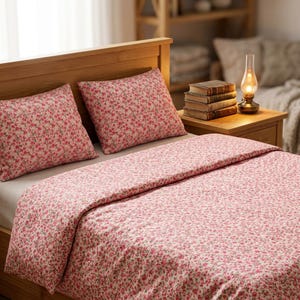 May include: A bedding set featuring a light pink fabric with a dense pattern of small red roses and green leaves. The set includes a large comforter and two matching rectangular pillows, all displayed on a wooden bed frame with a headboard.