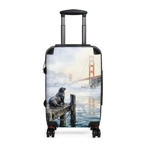 Może przedstawiać: Walizka na kółkach ze zdjęciem mostu Golden Gate w San Francisco. Lew morski siedzi na drewnianym molo. Most jest w kolorze czerwonopomarańczowym, na tle mglistego tła.