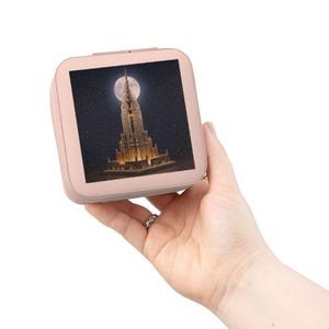 May include: A rose gold square jewellery box displaying an image of a tall building with a spire, lit against a night sky with a full moon. The box is held in a hand, highlighting its compact size and design.