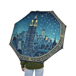 Könnte beinhalten: Ein offener Regenschirm mit einer nächtlichen Skyline von New York City, einschließlich des Empire State Building und des World Trade Centers. Der Schirm hat einen dunkelblauen Hintergrund mit weißen Sternen und einem dekorativen Goldrand.