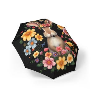 May include: Black umbrella featuring a watercolor illustration of a brown rabbit surrounded by colorful flowers. The rabbit wears a floral crown. The flowers include yellow, pink, blue, and orange blooms. The umbrella is open and ready for use.