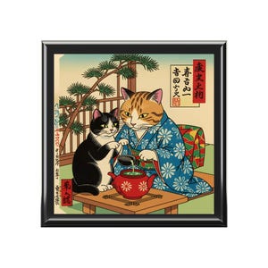 May include: A framed print depicting two cats in traditional Japanese clothing. One cat pours tea from a red teapot. The print includes Japanese text and is set within a black frame.