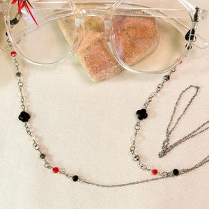 Könnte beinhalten: Brille mit transparentem Rahmen und silberner Brillenkettenhalterung. Die Kette hat schwarze, rote und klare Perlen sowie schwarze, blütenförmige Anhänger. Die Brille liegt auf einem kleinen, hellbraunen Stein.