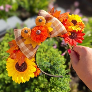 May include: A headband featuring a central beige and brown plaid bow, decorated with small orange pumpkins, yellow and red sunflowers, autumn-coloured fabric leaves, and small red berries. The headband has a thin, dark, flexible frame.