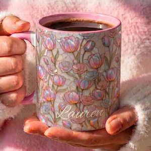 May include: A ceramic mug with a pink interior and handle, featuring an iridescent, stained-glass style pattern of pink and blue tulips. The name Lauren is written in white cursive script across the lower front of the mug, which is filled with dark coffee.
