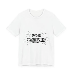 Könnte beinhalten: Weißes T-Shirt mit dem schwarzen Schriftzug "UNDER CONSTRUCTION", umgeben von dekorativen Linien. Das T-Shirt hat kurze Ärmel und einen Rundhalsausschnitt. Das Design ist schlicht und auffällig.