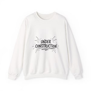 Könnte beinhalten: Weißes Sweatshirt mit Rundhalsausschnitt und dem schwarzen Schriftzug "UNDER CONSTRUCTION". Der Text ist stilisiert und von dekorativen Linien umgeben. Das Sweatshirt hat lange Ärmel und ein schlichtes, lässiges Design.