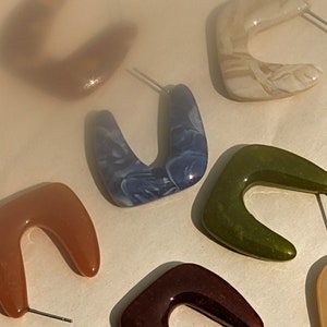 May include: A set of six colorful, geometric-shaped earrings. The earrings are made of resin and have a marbled effect. The earrings are in various colors, including brown, green, blue, and white.