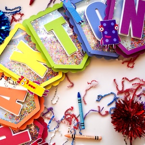 May include: A vibrant banner spelling "THANKS" with multi-coloured letters, each framed with a clear window filled with confetti. The image includes crayons, pipe cleaners, and decorative pom-poms, creating a festive and celebratory theme.
