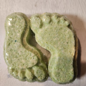 Puede incluir: Dos bombas de ba&ntilde;o verdes con forma de pies. Las bombas de ba&ntilde;o est&aacute;n cubiertas de motas blancas y tienen un tono verde claro.