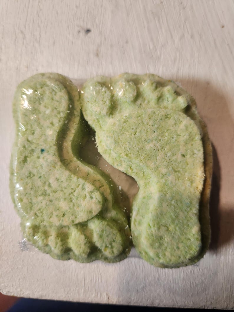 Puede incluir: Dos bombas de ba&ntilde;o verdes con forma de pies. Las bombas de ba&ntilde;o est&aacute;n cubiertas de motas blancas y tienen un tono verde claro.