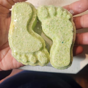 Puede incluir: Dos bombas de ba&ntilde;o verdes con forma de huellas. Las bombas de ba&ntilde;o est&aacute;n en una envoltura de pl&aacute;stico transparente.