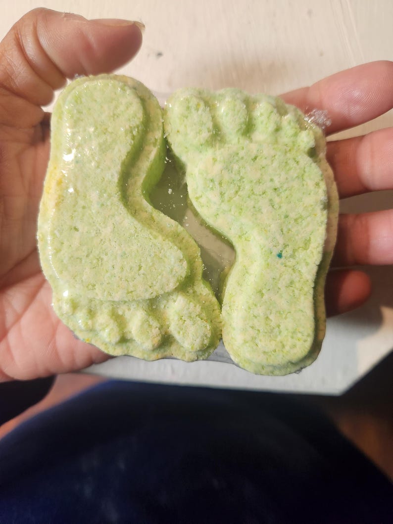 Puede incluir: Dos bombas de ba&ntilde;o verdes con forma de huellas. Las bombas de ba&ntilde;o est&aacute;n en una envoltura de pl&aacute;stico transparente.