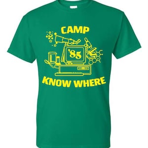 May include: Green t-shirt with a yellow graphic design. The design includes the text "CAMP KNOW WHERE '85" along with illustrations of a computer, microscope, and other science-related elements. The shirt is a short-sleeved crew neck.