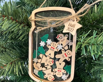Puede incluir: Un adorno navideño de madera con forma de tarro de cristal con una ventana transparente. El tarro está lleno de figuras de pan de jengibre con nombres escritos en ellas, incluyendo "Mom", "Dad", "Wade", "Laci", "Sage" y "June". El adorno también incluye una pequeña estrella con el texto "Hull 2022" en ella.
