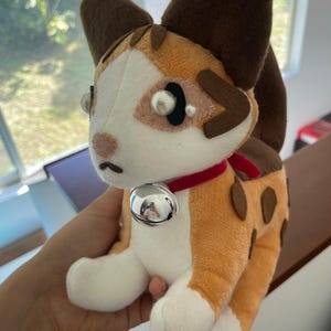 May include: A plush cat toy with a white face, orange and brown spots, and large brown ears. It has a red collar with a silver bell. The toy is made of soft fabric and has a cute, cartoonish design.