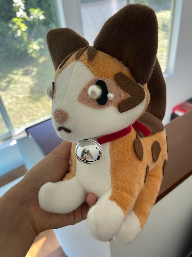 May include: A plush cat toy with a white face, orange and brown spots, and large brown ears. It has a red collar with a silver bell. The toy is made of soft fabric and has a cute, cartoonish design.