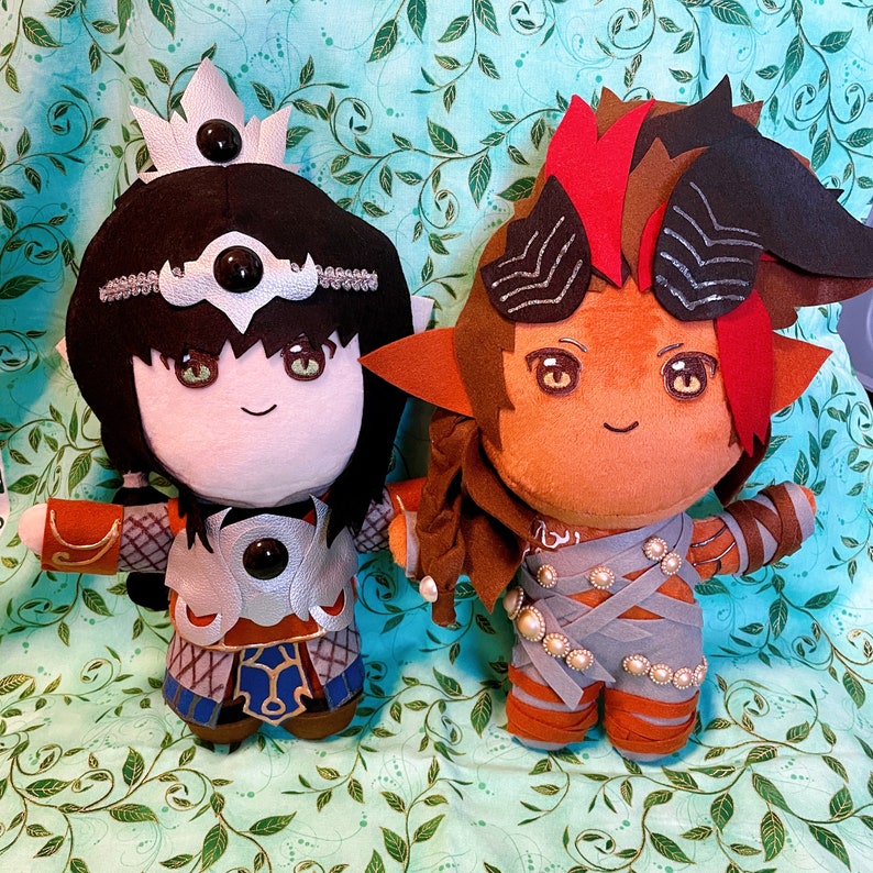 May include: Two plush dolls with detailed costumes. One doll has black hair, a crown, and silver armour. The other doll has orange fur, horns, and wrapped grey fabric with pearl accents. Both dolls have embroidered facial features.