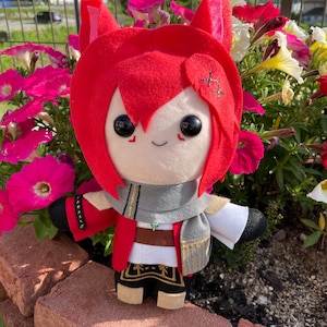 May include: A plush doll with red hair and cat ears, wearing a red jacket, gray scarf, and black pants with gold trim. The doll has large black eyes and is standing in front of pink and white flowers.