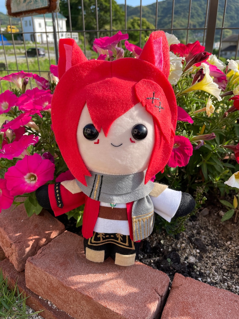 May include: A plush doll with red hair and cat ears, wearing a red jacket, gray scarf, and black pants with gold trim. The doll has large black eyes and is standing in front of pink and white flowers.