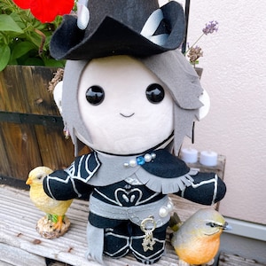 Puede incluir: Una muñeca de peluche con cabello gris que lleva un sombrero negro y un atuendo negro y gris con detalles plateados. La muñeca tiene ojos negros grandes, una pequeña sonrisa bordada y un dije de estrella. Está flanqueada por un pollito amarillo y un pájaro.