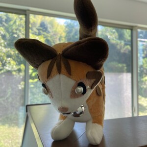 May include: A plush cat toy with a tan and white body, brown ears, and a matching tail. The cat has a silver bell collar and pearl-like accents around the eyes. The toy is standing on a brown surface.