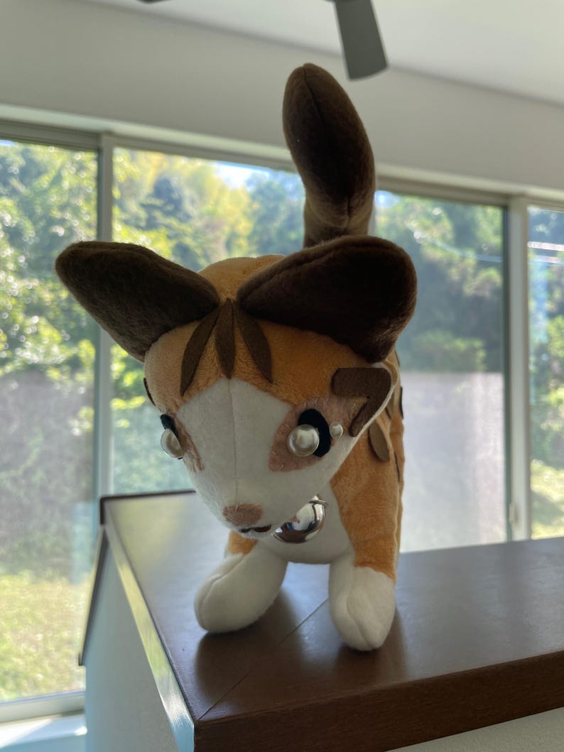 May include: A plush cat toy with a tan and white body, brown ears, and a matching tail. The cat has a silver bell collar and pearl-like accents around the eyes. The toy is standing on a brown surface.