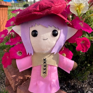 May include: A handmade plush doll with a whimsical design. The doll features a pink dress, a tan vest with black stitching, and a burgundy beret. It has large black eyes, lavender hair, and brown arms.