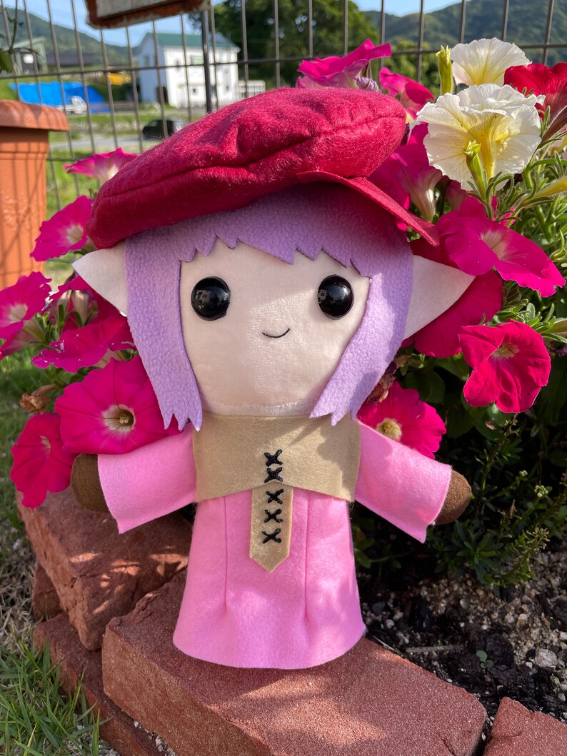 May include: A handmade plush doll with a whimsical design. The doll features a pink dress, a tan vest with black stitching, and a burgundy beret. It has large black eyes, lavender hair, and brown arms.
