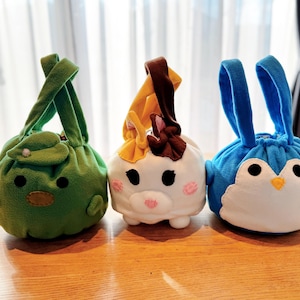 Puede incluir: Tres bolsas de felpa con cordón en forma de animales. Una bolsa de rana verde, una bolsa de gato blanco con mejillas rosadas y una bolsa de pingüino azul. Cada bolsa tiene un cierre de cordón y un asa.