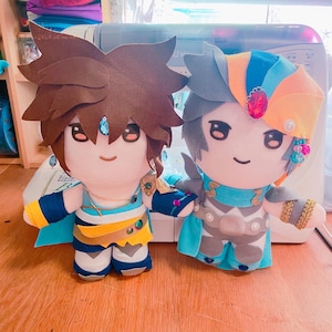 Könnte beinhalten: Zwei Plüschtiere, eines mit braunen Haaren und einem blauen und gelben Outfit, das andere mit grauen Haaren und einem blauen und weißen Outfit. Beide Spielzeuge haben große Augen und lächelnde Gesichter.