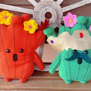 May include: Two plush cactus and tree-like figures with felt flowers. The orange figure has yellow flowers, while the green figure has pink flowers and a felt moustache. They are sitting on a wooden bench.