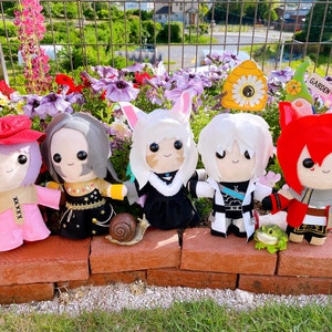 Puede incluir: Cinco muñecos de peluche de estilo anime, cada uno con atuendos y peinados únicos, están colocados en una repisa de ladrillo frente a un parterre de flores. Los muñecos tienen varios colores, incluyendo rosa, rojo, negro y blanco. También hay una figura de caracol y una pequeña rana.