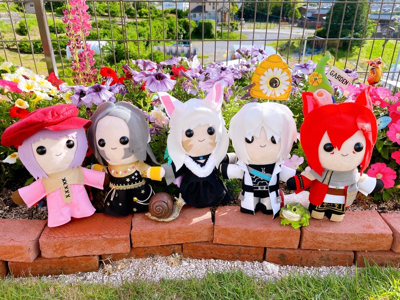 May include: Five plush dolls with anime-style features, each with unique outfits and hairstyles, are arranged on a brick ledge in front of a flower bed. The dolls have various colors, including pink, red, black, and white. A snail figurine and a small frog are also present.