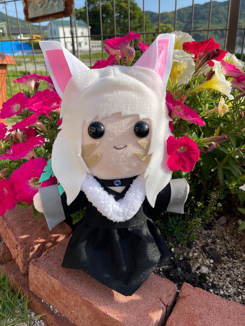 May include: A plush doll with white fur, pink-lined ears, and large black eyes. The doll wears a black dress with a white fur collar and a blue gem. The doll is set against a backdrop of vibrant pink and white flowers.