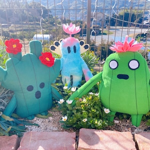 May include: Three plush toys in a garden. A green cactus toy with red flower details, a blue figure with wings, and a green cactus toy with a pink flower. The toys are set amongst flowers and greenery, with a fence and hills in the background.