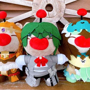 May include: Three plush dolls with large red noses and round red toppers. The dolls are dressed in various outfits, including a gold and brown ensemble, a grey suit with a red bow tie, and a teal and brown dress. They are set against a wooden background.