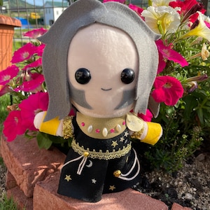 May include: A plush doll with a light skin tone, gray hair, and black eyes. The doll wears a black outfit with gold accents, stars, and a crescent moon design. The doll is posed in front of pink and white flowers.