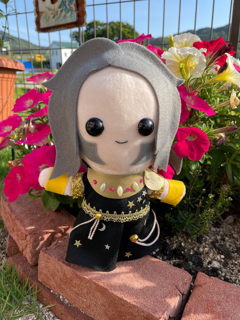 May include: A plush doll with a light skin tone, gray hair, and black eyes. The doll wears a black outfit with gold accents, stars, and a crescent moon design. The doll is posed in front of pink and white flowers.