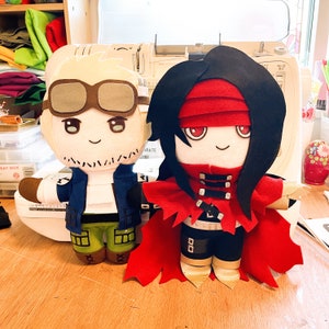 May include: Two handmade plush dolls. One doll has blonde hair, brown goggles, and a blue jacket. The other doll has long black hair, a red cape, and a red headband. Both dolls have smiling faces and are made of felt.