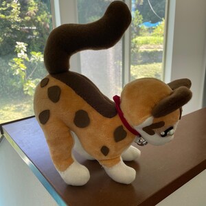 May include: A plush cat toy in a playful pose. The cat has a tan body with brown spots and a brown tail. It wears a red collar with a silver bell. The cat's paws and face are white.