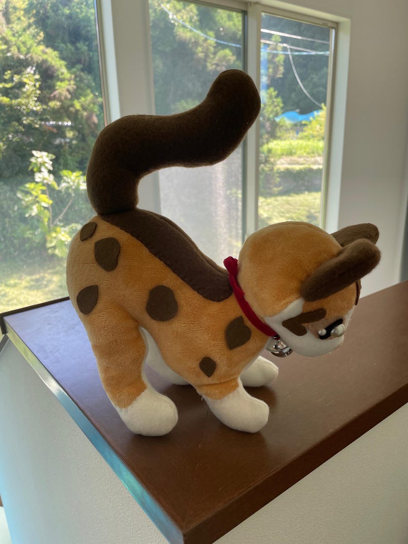 May include: A plush cat toy in a playful pose. The cat has a tan body with brown spots and a brown tail. It wears a red collar with a silver bell. The cat's paws and face are white.