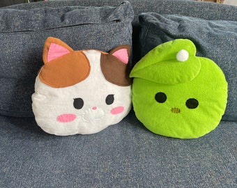 Könnte beinhalten: Zwei Plüschkissen: eines in Form einer Katze mit weißen, braunen und rosa Details und das andere in Form eines grünen Apfels mit einem Blatt und einem weißen Bommel. Beide Kissen haben schwarze Augen und liegen auf einem blauen Sofa.