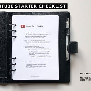 Puede incluir: Una agenda negra con una página blanca abierta en una lista de verificación titulada "Youtube Starter Checklist". La lista de verificación incluye pasos para crear un canal de YouTube, verificar un programa de socios de canal, crear una pantalla final personalizada y crear una marca para tu canal. La agenda tiene un archivador de anillas negro y un bolígrafo blanco sobresale de la parte superior.