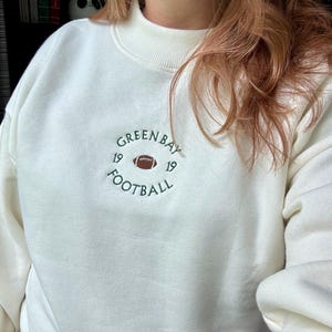 May include: A cream-colored crewneck sweatshirt featuring dark green embroidered text that reads GREEN BAY FOOTBALL in a circular pattern. In the center, there is a small brown football icon flanked by the number 19 on both the left and right sides.
