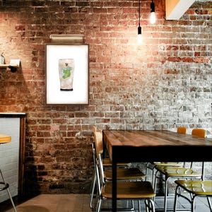 May include: A framed watercolor painting of a pint glass filled with a green liquid. The glass is on a white background. The painting is hanging on a brick wall in a restaurant setting.