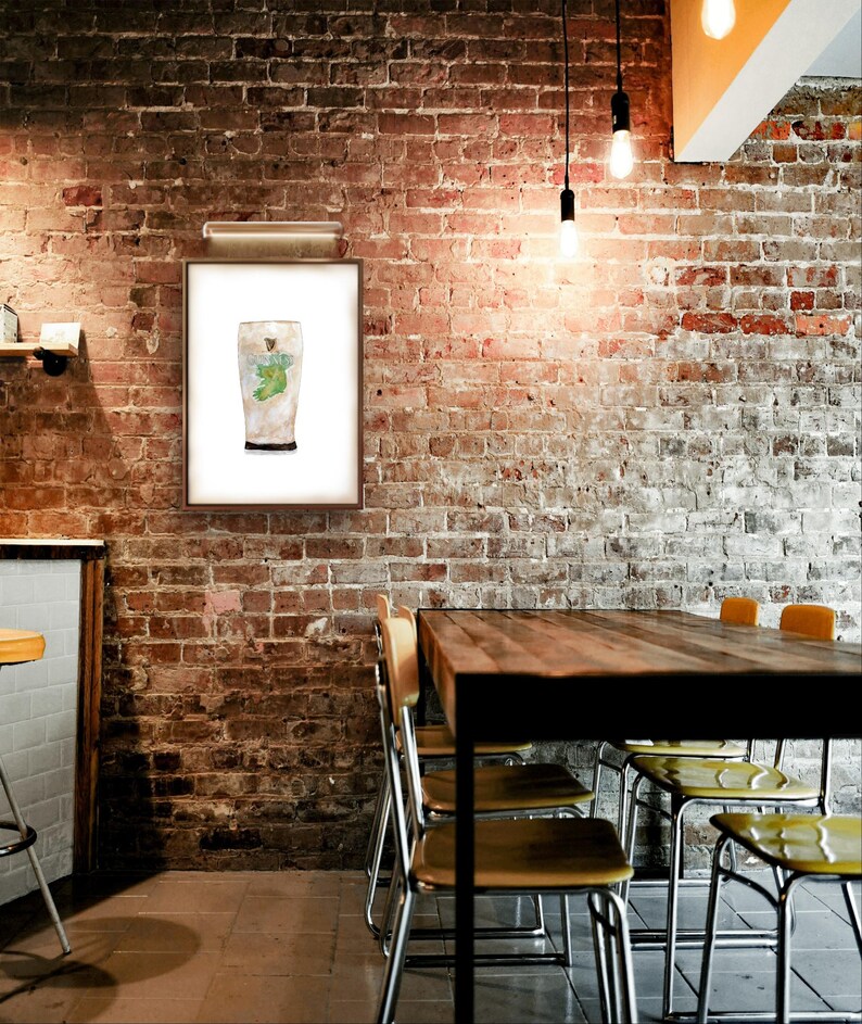 May include: A framed watercolor painting of a pint glass filled with a green liquid. The glass is on a white background. The painting is hanging on a brick wall in a restaurant setting.